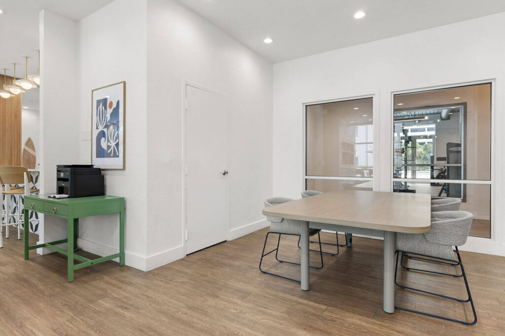 Modern office space with a wooden table, six gray chairs, a green side table with a printer, a framed artwork on the wall, and two large interior windows. The floor is wood and lighting is recessed.