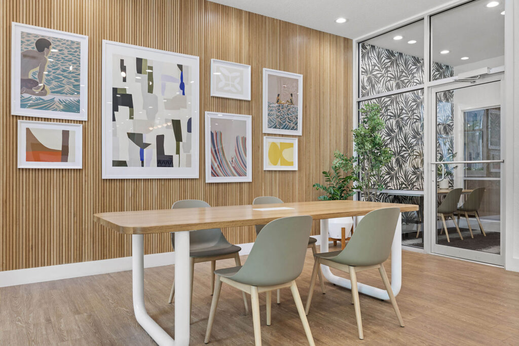 A modern meeting room with a wooden table, four gray chairs, framed abstract art on a wood-paneled wall, a glass door, potted plant, and a patterned accent wall.