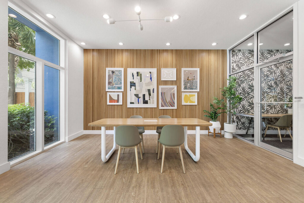 Modern conference room with wood floors, a wooden table, four green chairs, abstract art on a wood-paneled wall, large windows, and a glass wall showing a room with bold leaf-patterned wallpaper and potted plants.