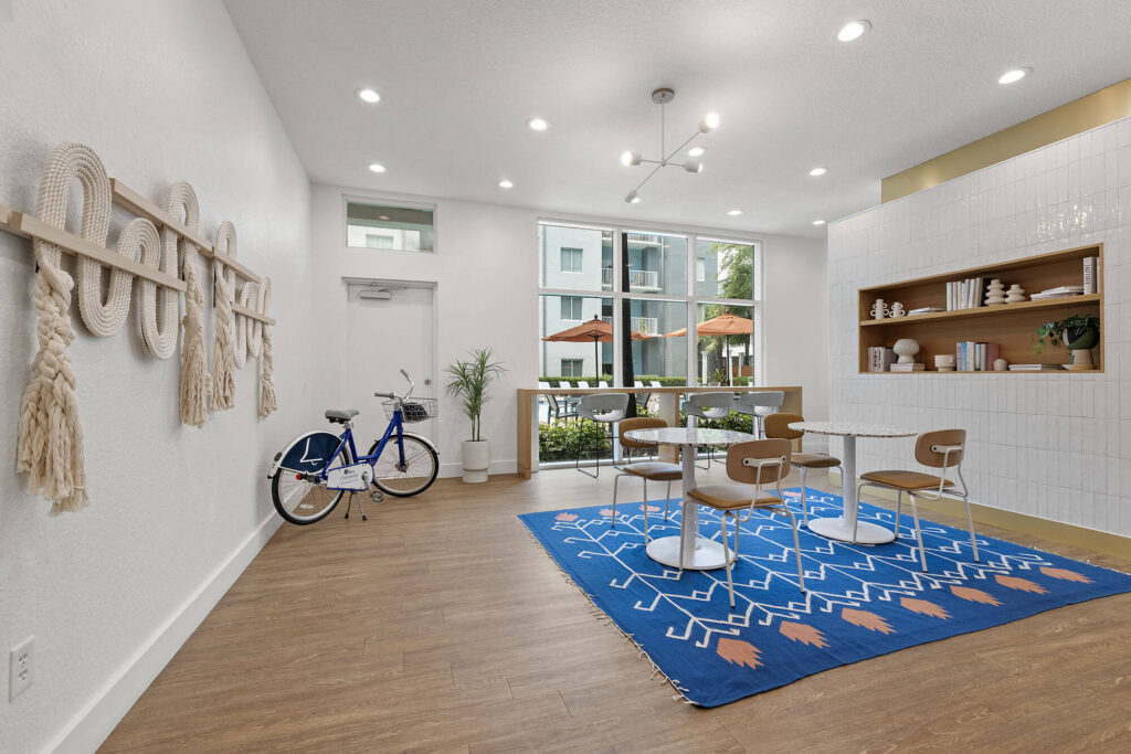 Modern, bright lounge with white walls, wooden floor, blue geometric rug, small tables and chairs, wall shelves with decor, a bicycle, macrame wall hangings, and large windows showing outdoor umbrellas.