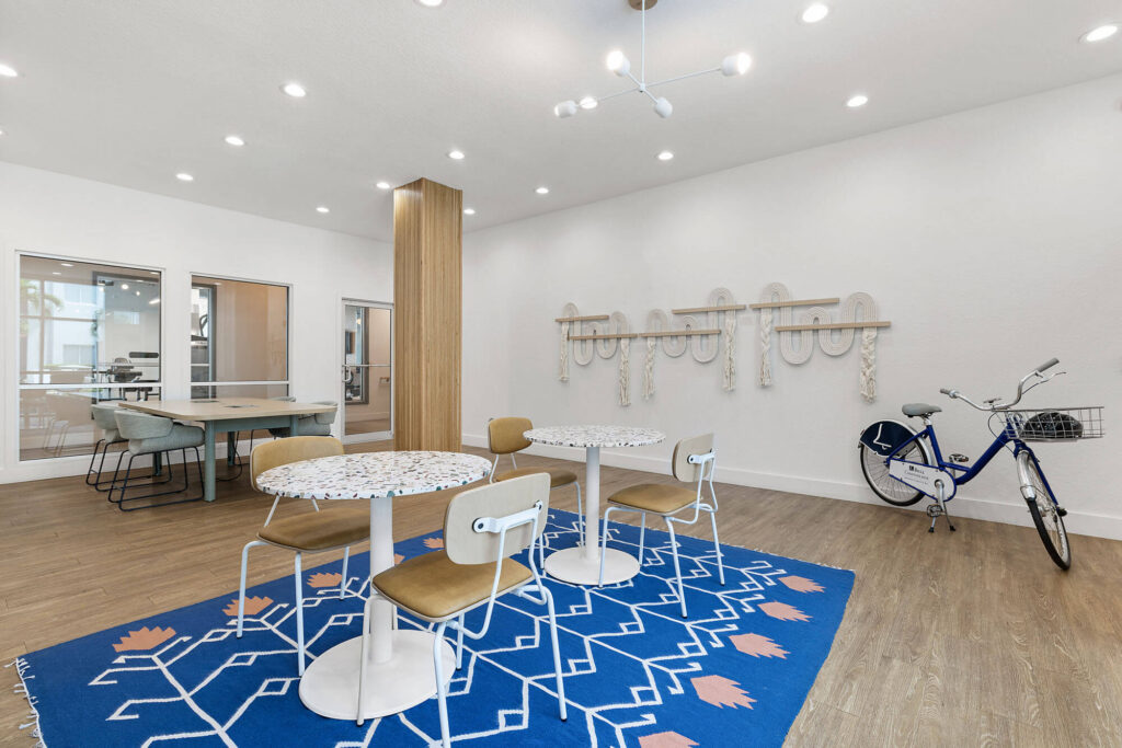 Modern room with three round tables and chairs on a blue rug, a bike against the wall, coat hooks with ropes, wooden floors, and an adjacent glass-walled meeting area in the background.