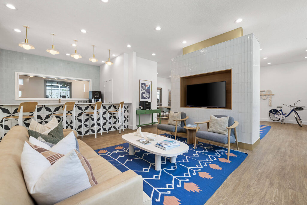 Modern lounge with a tan sofa, two gray chairs, a blue patterned rug, a coffee table with books, a large TV, a bicycle, and a bar area with geometric stools under gold pendant lights.