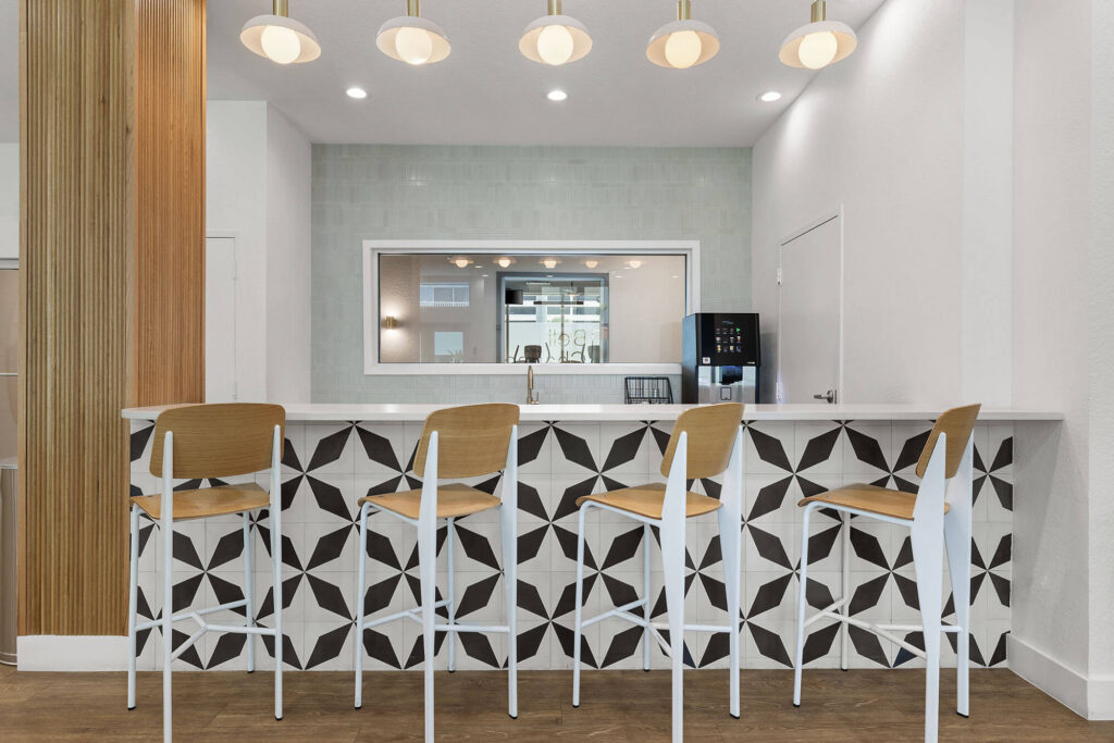 Modern cafe counter with geometric black-and-white tile, three wooden bar stools with white metal legs, pendant lights above, a large window at the back, and a coffee machine on the right side of the counter.