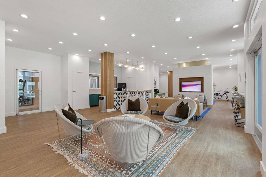 Modern lounge area with white walls, wood flooring, round chairs, decorative rug, large TV, and geometric reception desk. Bright lighting, glass doors, and a bicycle in the background give the space a sleek, welcoming feel.