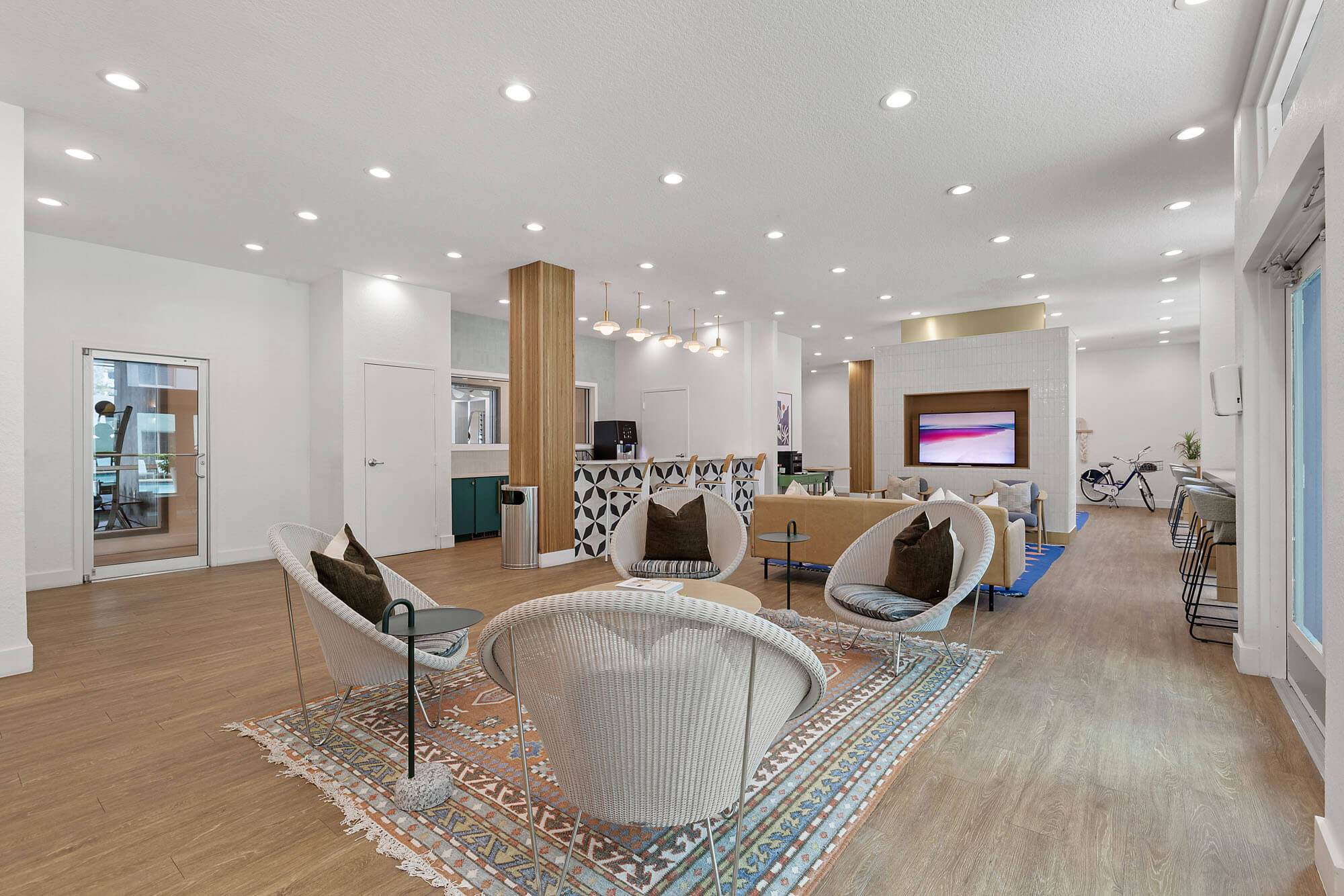 Modern lounge area with white walls, wood flooring, round chairs, decorative rug, large TV, and geometric reception desk. Bright lighting, glass doors, and a bicycle in the background give the space a sleek, welcoming feel.