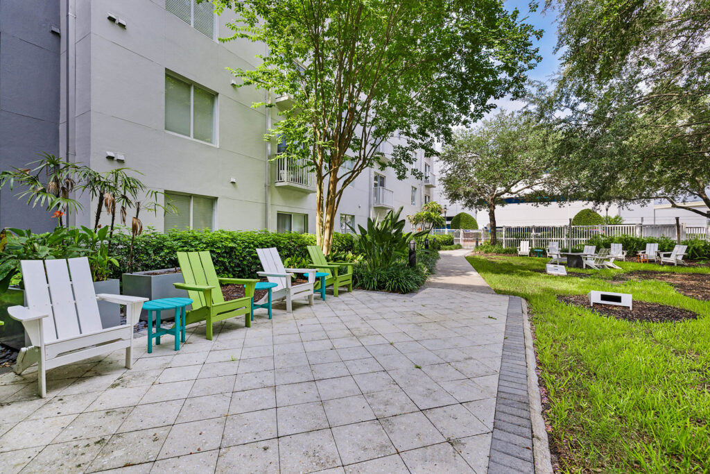 Outdoor courtyard with white and green chairs, small tables, trees, and lush plants beside a modern apartment building on a sunny day. Pathways and additional seating are visible in the background.
