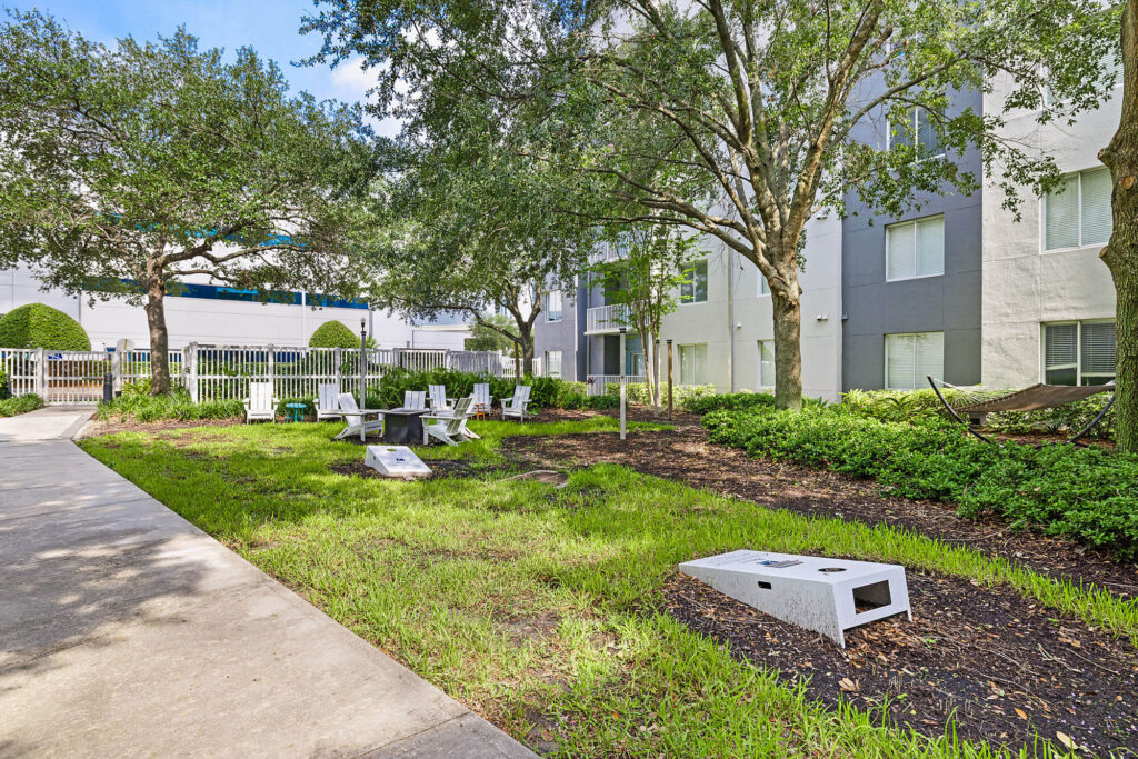 A courtyard features Adirondack chairs around a firepit, a hammock between trees, and a cornhole game set on the grass, with a modern apartment building in the background.