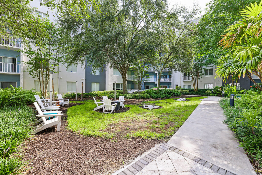 A landscaped courtyard with white Adirondack chairs arranged around a fire pit, surrounded by green grass, trees, and apartment buildings in the background. A paved walkway runs alongside the seating area.