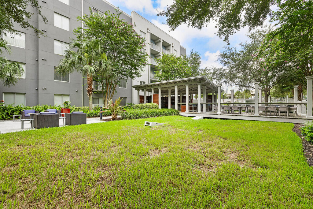 A grassy courtyard with patio seating, palm trees, and a modern gray apartment building in the background. A covered outdoor seating area is on the right side.