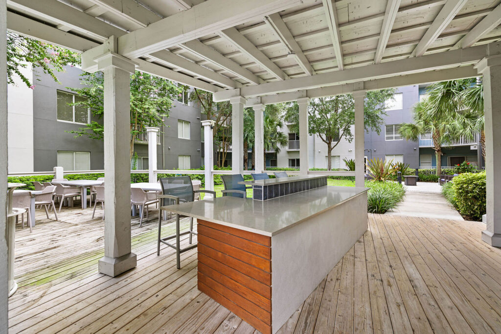 Covered outdoor patio with a large countertop island, barstools, wood flooring, and dining tables, surrounded by greenery and apartment buildings in the background.