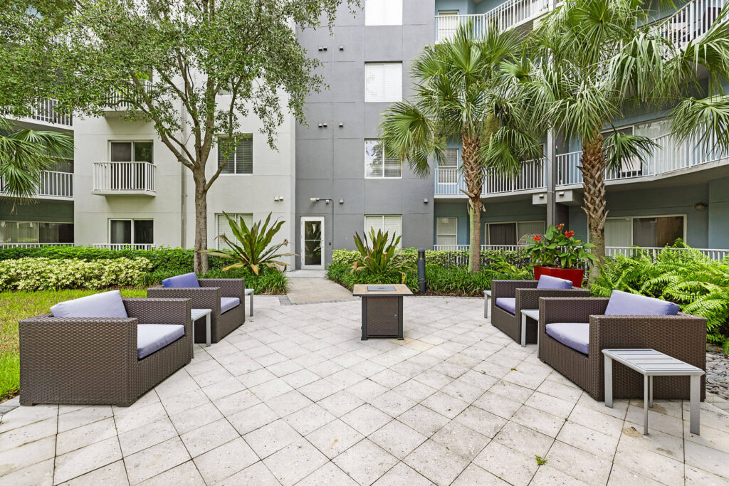 Outdoor patio with wicker seating, gray cushions, a fire pit, side tables, and lush landscaping. Apartment balconies are in the background, with palm trees and other plants surrounding the seating area.