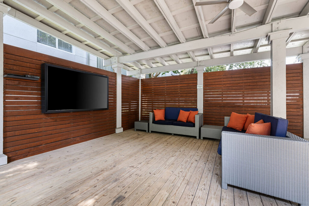 Modern covered patio with wooden floor and slatted wood walls, featuring two wicker sofas with orange and navy cushions, and a large flat-screen TV mounted on the wall.