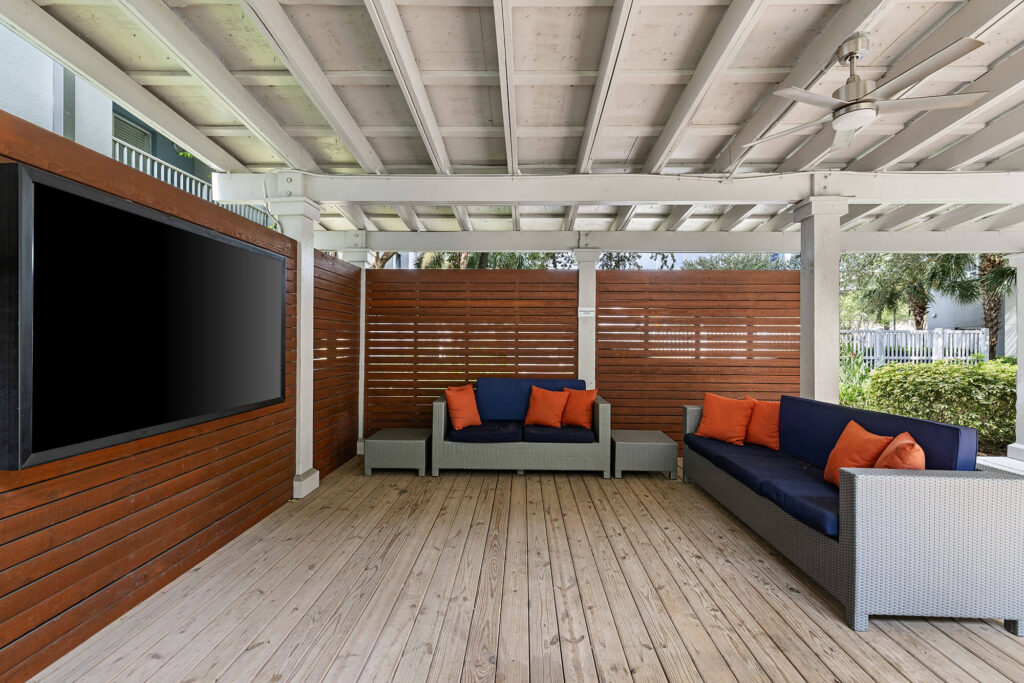 Covered outdoor lounge area with wooden floor, two wicker sofas with blue cushions and orange pillows, a large flat-screen TV on a wooden wall, ceiling fan, and partial sunlight.