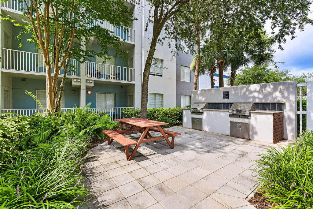 A wooden picnic table sits on a patio near two stainless steel grills. Green plants and trees surround the area, with a white apartment building visible in the background.