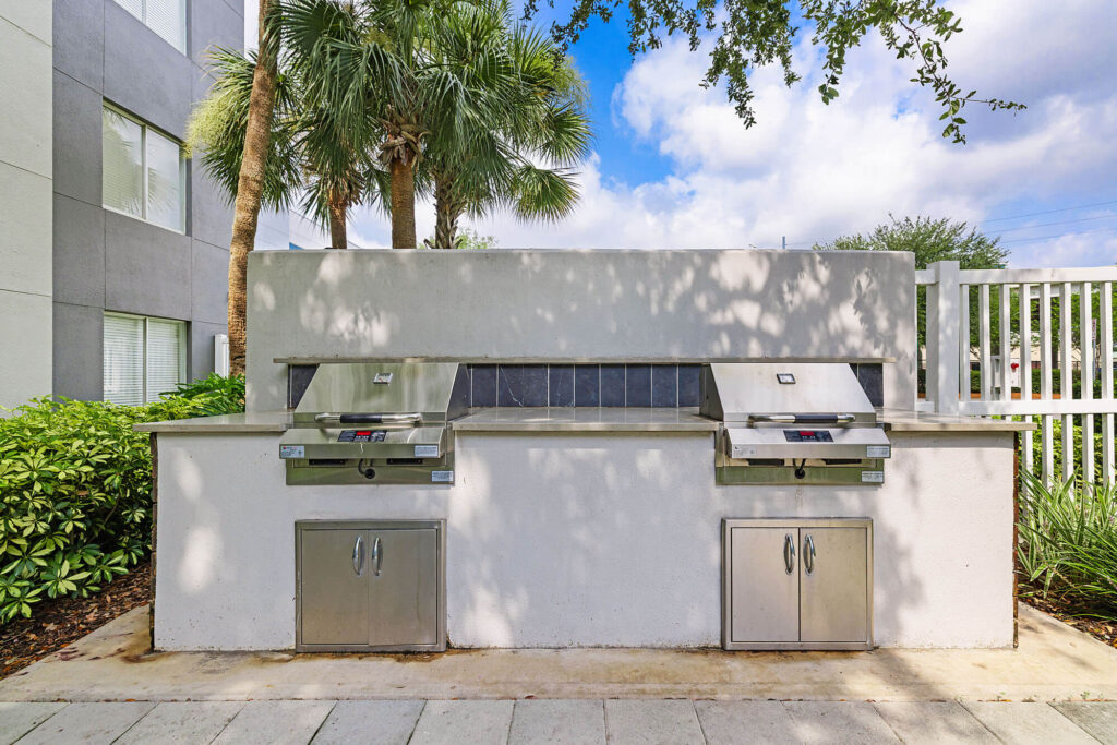 Outdoor grilling area with two stainless steel grills, storage cabinets below, and palm trees in the background next to a white fence and building.