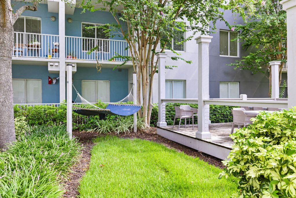 A blue hammock hangs between two posts near green grass and shrubs, with a patio area featuring tables and chairs, in front of a modern apartment building.