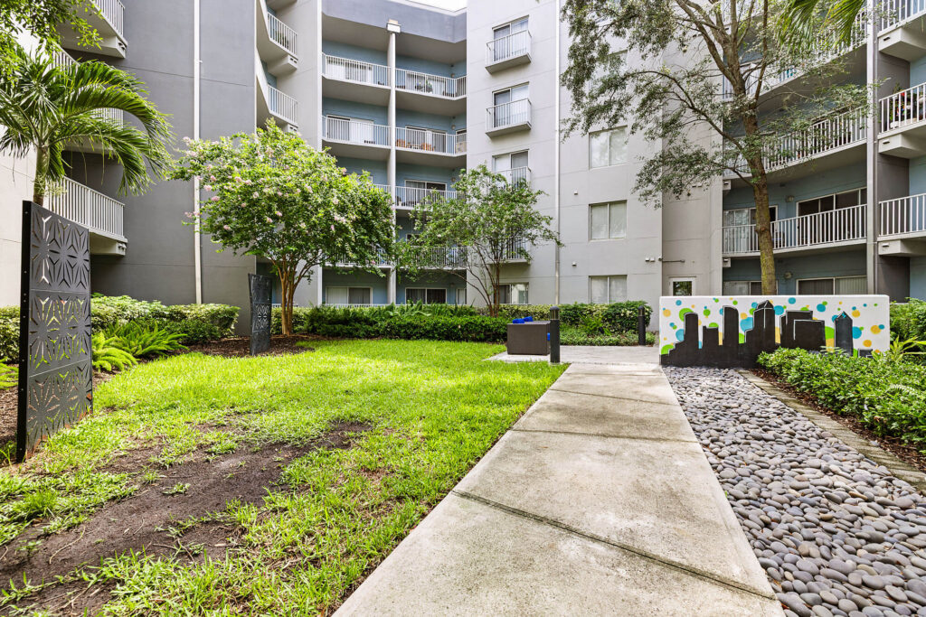 A courtyard in an apartment complex with green grass, trees, a walkway, decorative rocks, and a painted mural on the right. Gray balconies and windows line the surrounding buildings.