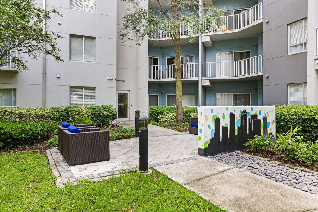 A modern apartment courtyard with outdoor seating, greenery, a mural featuring city buildings, balconies, and a paved walkway.