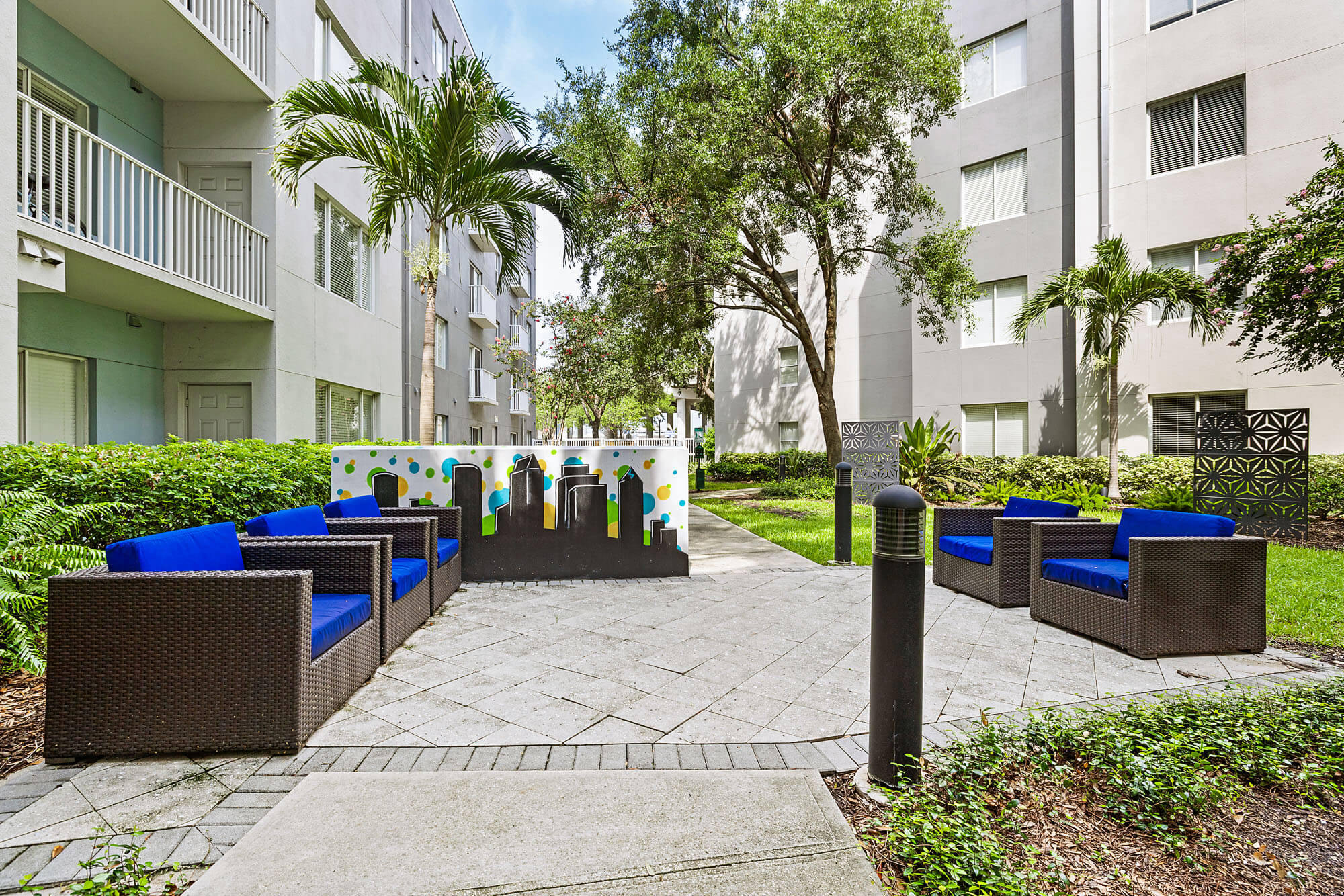 Outdoor courtyard with four blue-cushioned wicker sofas arranged around a modern mural, surrounded by greenery and trees, with gray apartment buildings in the background.