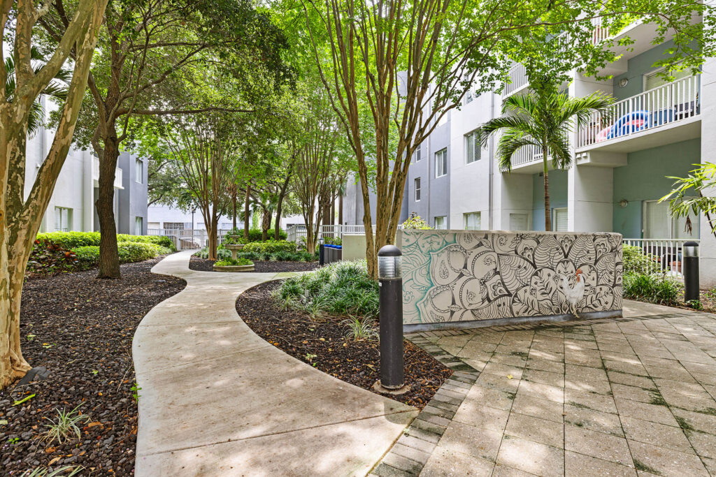 Curved sidewalk and tiled pathway winding through landscaped greenery, trees, and shrubs beside modern white apartment buildings with balconies, featuring a wall decorated with artistic black and white sketches.