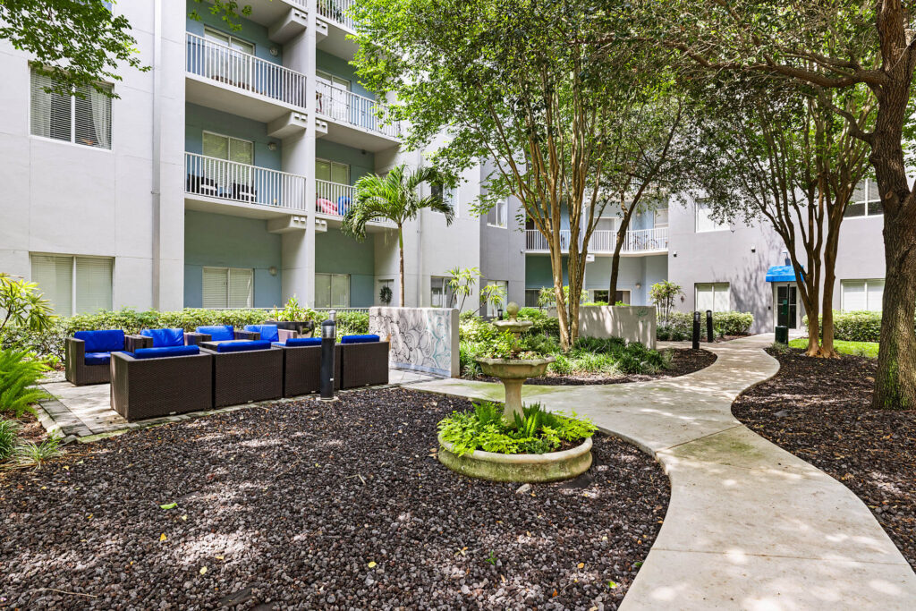 Curved walkway in an apartment courtyard with trees, a round birdbath, outdoor seating with blue cushions, and a three-story gray building in the background.