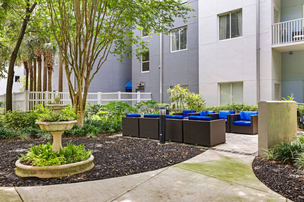 Outdoor courtyard with wicker patio seating featuring blue cushions, surrounded by greenery, trees, a birdbath, and a curved walkway next to a modern gray-and-white apartment building.