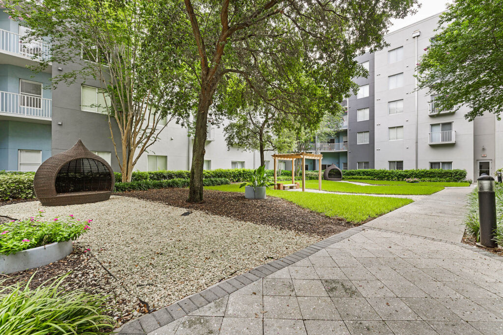 A modern apartment courtyard with trees, green grass, stone paths, a wicker lounge chair, a small wooden pergola, and landscaped garden beds near a gray multi-story building.