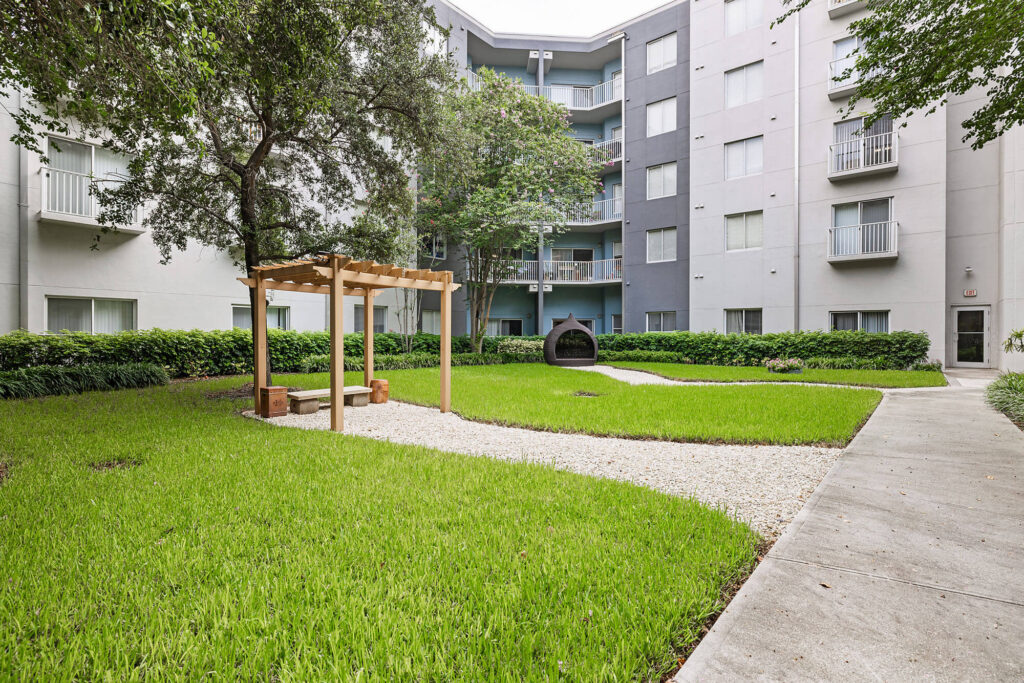 A courtyard with green grass, a gravel path, wooden pergola with benches, and a small arched structure, surrounded by a four-story apartment building and trees.