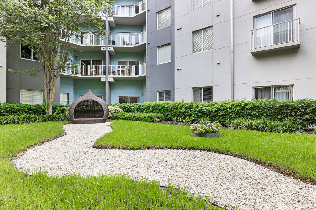 A modern apartment courtyard with green grass, a gravel walkway, a wicker seat under a tree, manicured hedges, and balconies overlooking the garden.