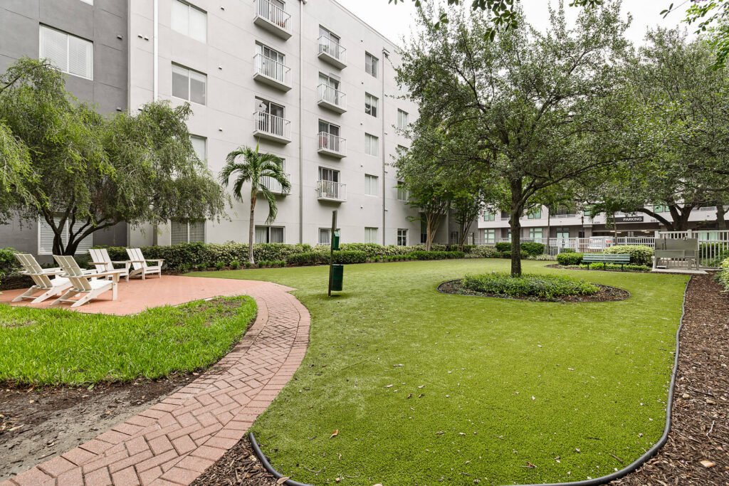A landscaped courtyard with green grass, trees, lounge chairs, a bench, and a curving brick pathway beside a multi-story apartment building.
