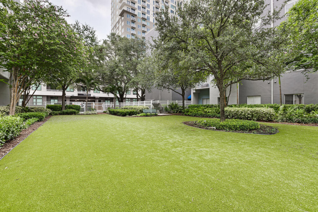 A well-maintained courtyard with artificial grass, surrounded by trees, bushes, benches, and modern apartment buildings in the background.
