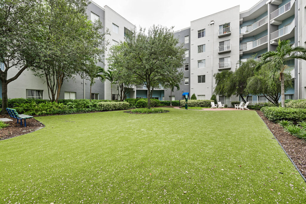 A green courtyard with artificial grass, trees, shrubs, and seating areas surrounded by multi-story apartment buildings with balconies and windows.