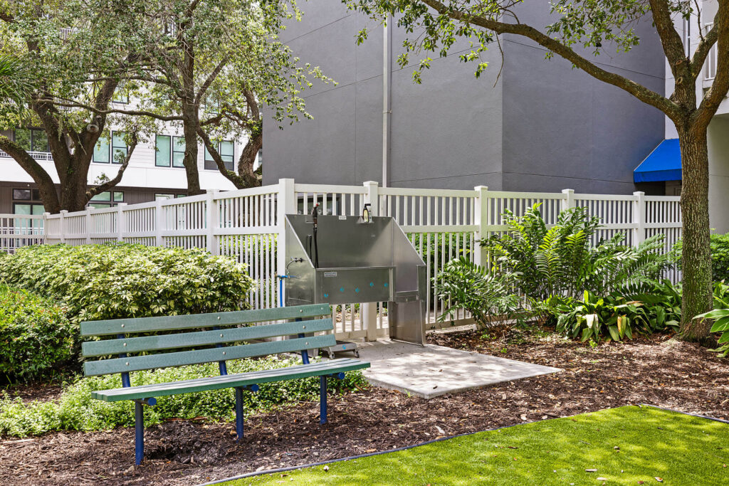 A green bench sits near a fenced area with lush plants and trees. Behind the bench, there is a stainless steel dog washing station on a concrete pad.