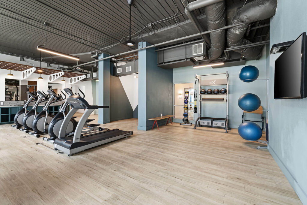 Modern gym with treadmills, exercise balls, weight rack, wall-mounted TV, light wood flooring, and exposed ceiling pipes. The space is clean, open, and well-lit with light blue and gray walls.