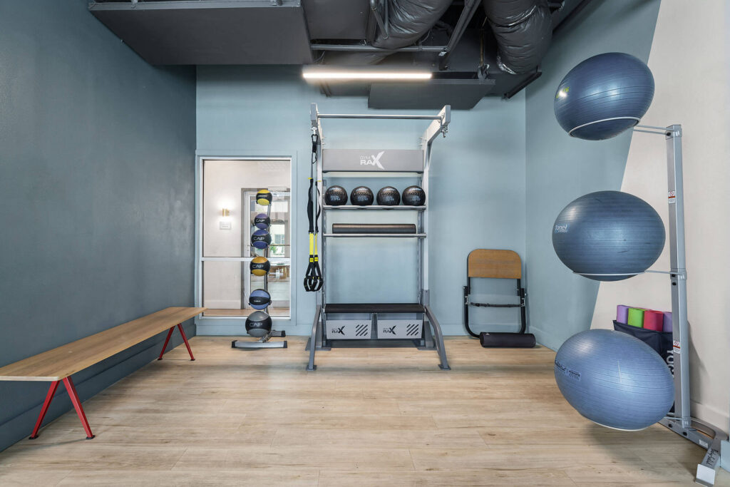 Small fitness room with exercise balls on a rack, medicine balls on shelves, resistance bands, a wooden bench, and a chair against blue walls and light wood flooring. Ceiling features exposed black ductwork.