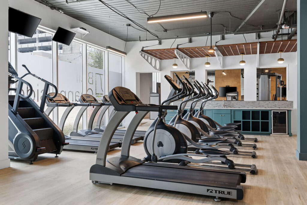 Modern gym with treadmills, stationary bikes, a stair climber, large mirrors, hanging lights, and big windows letting in natural light. Light wood flooring and teal accents are visible throughout the space.