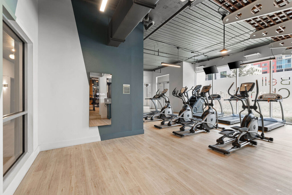 A modern gym with four elliptical machines on wood flooring, large windows letting in natural light, a mirror on a gray accent wall, and ceiling lights.