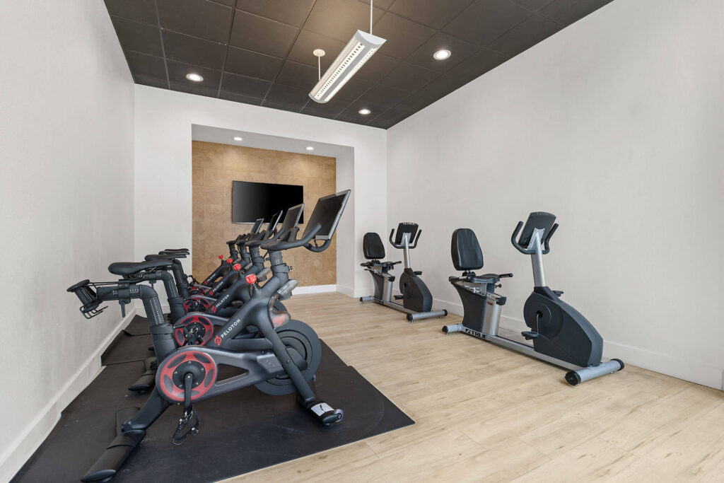 A small gym room with four stationary bikes on the left and two recumbent bikes on the right, wood flooring, white walls, and a mounted TV at the far end.