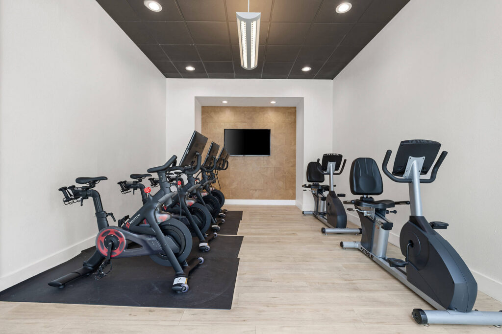 A small gym room with five stationary bikes on the left, two recumbent bikes on the right, light wood flooring, white walls, and a wall-mounted TV at the far end.