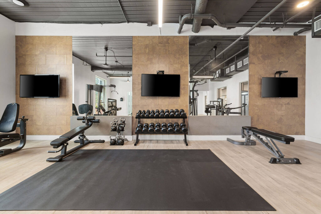 Modern gym with benches, dumbbells, kettlebells, three wall-mounted TVs, large mirrors, and exercise equipment. The floor has a black mat and light wood, with neutral-colored walls and ceiling pipes visible.