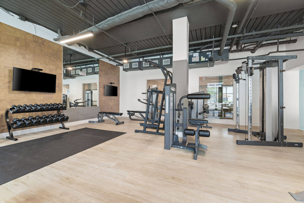 Modern gym with wood flooring, free weights, benches, exercise machines, and wall-mounted TVs. Large windows bring in natural light. Black ceiling with exposed ducts and mirrors reflecting the space.