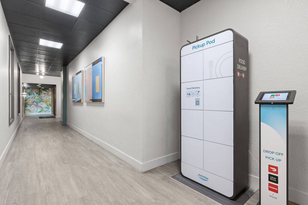 A white food delivery locker labeled Pickup Pod stands in a hallway beside a touchscreen kiosk. The corridor has wood flooring, wall art, and bright lighting.