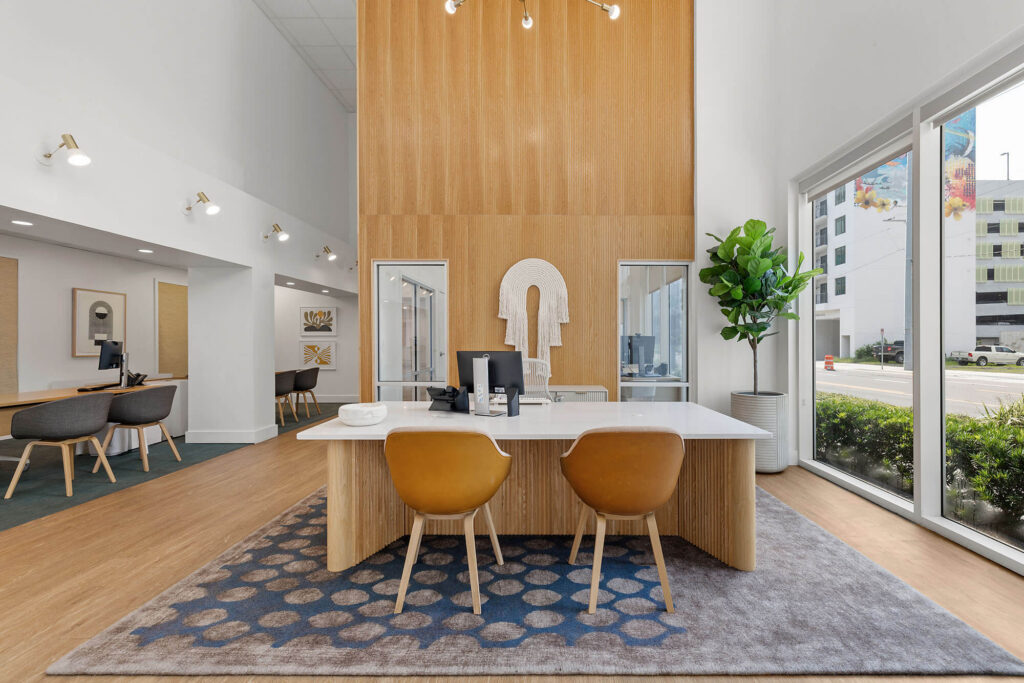 Modern office with a wooden accent wall, desks with computers, tan chairs, decorative plant, large windows, and city view outside. Contemporary design with natural light and clean lines.