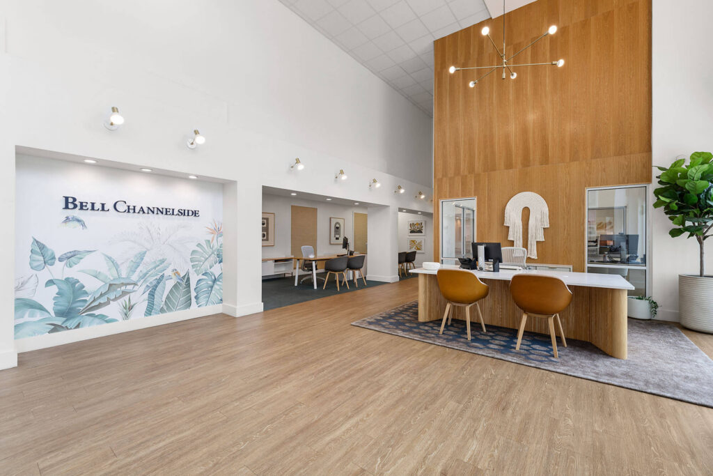 Modern office lobby with wooden floors, white walls, a mural reading Bell Channelside, and stylish furniture. Reception desk with two chairs is in front of a wood accent wall. Large plant is in the corner.