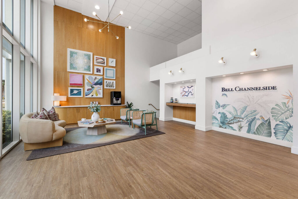 Modern lobby with wood flooring, high ceilings, a seating area with couches and chairs, wall art, large windows, and a reception desk labeled Bell Channelside. Bright, airy space with contemporary decor and indoor plants.