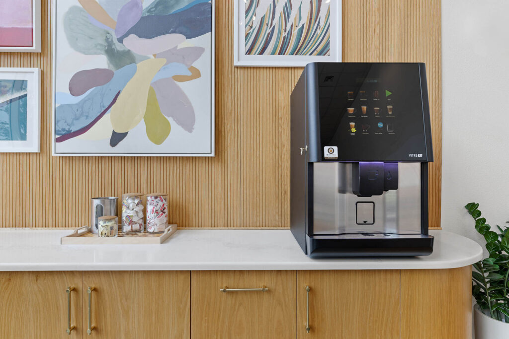 A modern coffee machine sits on a white countertop next to jars with stirrers and marshmallows. Colorful abstract art hangs on a wooden wall above the counter.