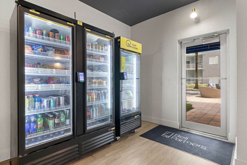 Two glass-door vending coolers filled with drinks and snacks stand in a bright room. A door leads outside to a courtyard. A dark mat by the door has the Bell Partners logo. A wall light is above the door.