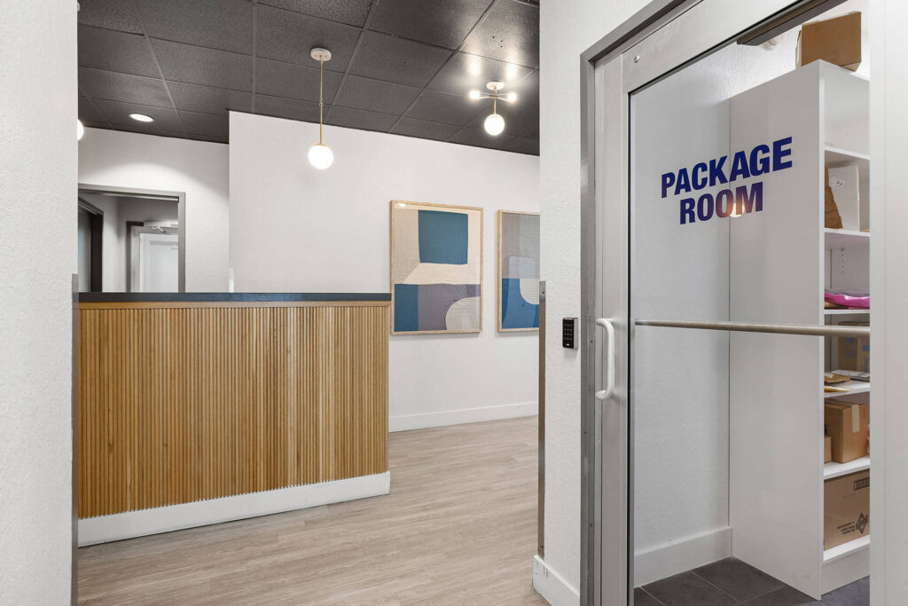 Modern lobby with a wood-paneled reception desk, abstract artwork on the wall, light wood flooring, and a glass door labeled Package Room showing shelves with packages inside.