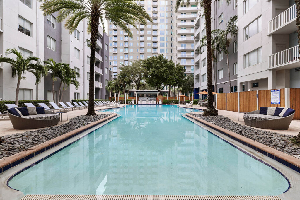A rectangular outdoor pool is surrounded by lounge chairs, palm trees, and modern apartment buildings, with a shaded seating area at the far end.