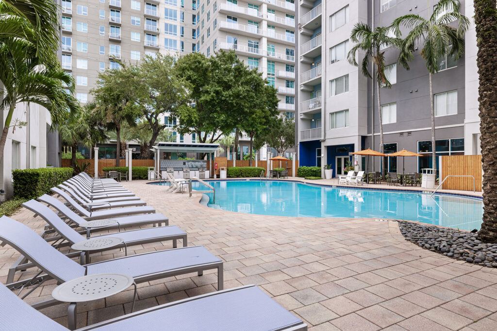 Outdoor pool area at a modern apartment complex with lounge chairs, tables, palm trees, and high-rise buildings in the background under a clear sky.